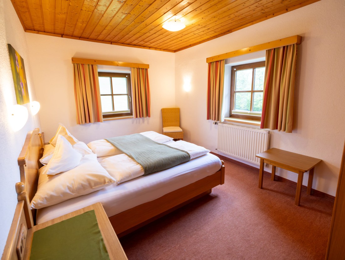 Schlicht eingerichtetes Schlafzimmer mit Holzmöbeln und zwei Fenstern mit Vorhängen