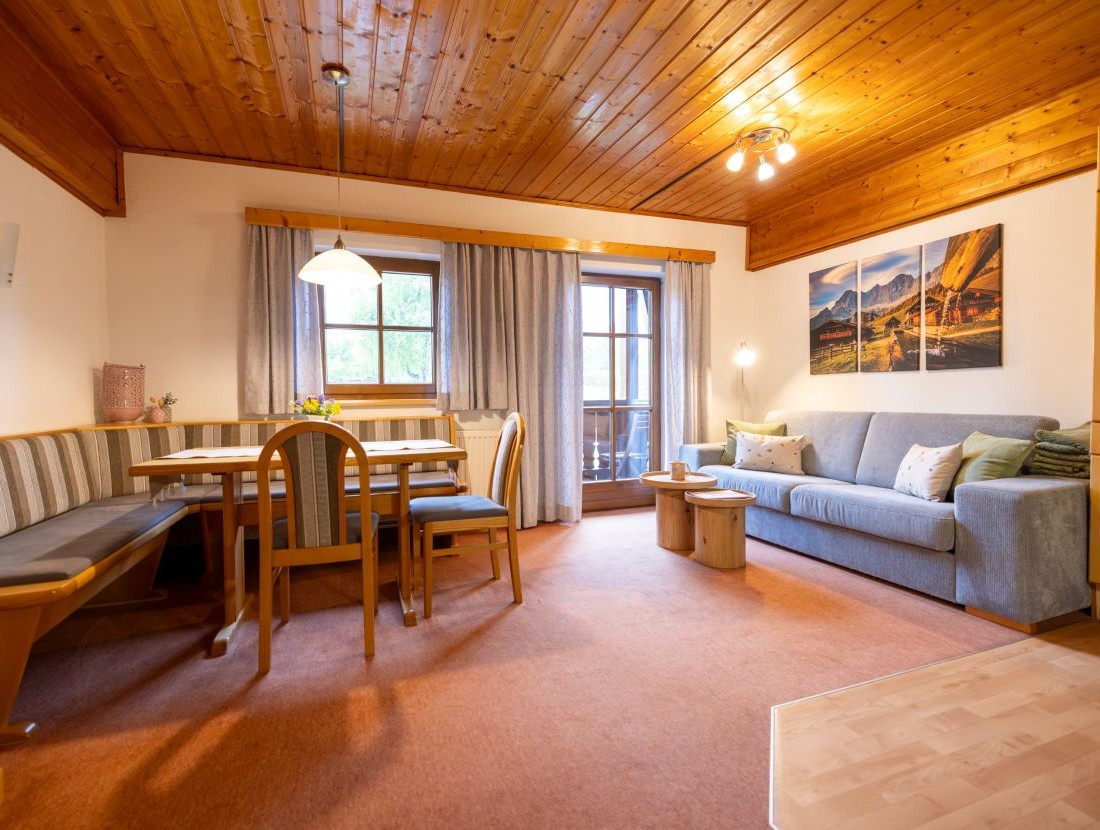 Wohnzimmer mit Holztäfelung, grauem Sofa, Esstisch und einem Balkon mit Vorhängen