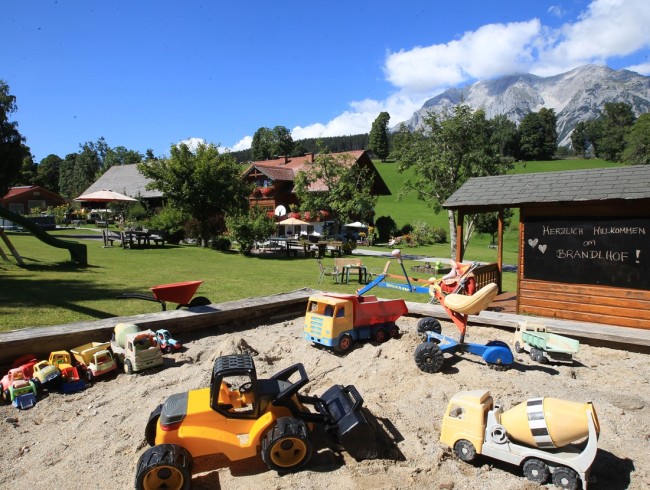 Spielzeuge im Sandkasten vor einer alpinen Landschaft mit Gebäuden