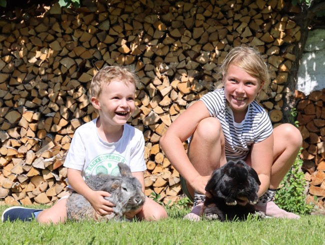 Zwei Kinder sitzen im Gras und halten Kaninchen vor einem Holzstapel